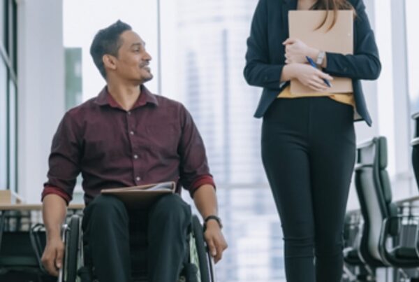 A man in a wheelchair is next to a walking woman in a business hallway